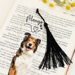 May include: A black and white bookmark with a tassel. The bookmark features a photo of a brown and white dog with the word "Mango" written in cursive. The bookmark also has a heart with paw prints and the text "The heart would do what he say, "This did I go for..."