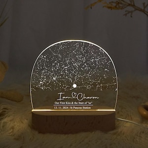 May include: A personalized night light featuring a constellation map. The clear acrylic panel displays a star chart with the names "Iam & Charon" and the date "13. 11. 2024". The light sits on a wooden base and emits a warm glow.
