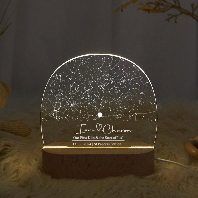 May include: A personalized night light featuring a constellation map. The clear acrylic panel displays a star chart with the names "Iam & Charon" and the date "13. 11. 2024". The light sits on a wooden base and emits a warm glow.