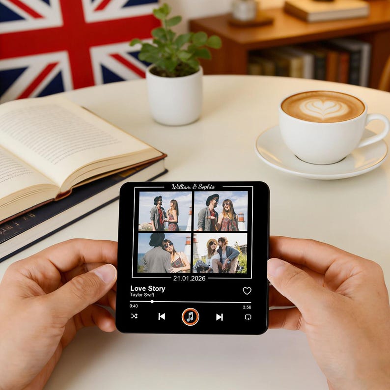 May include: A black smartphone screen shows a photo collage with the names "William & Sophia" and the date "21.01.2026". The screen displays a music player with the song "Love Story" by Taylor Swift. The phone is held in front of a white table.
