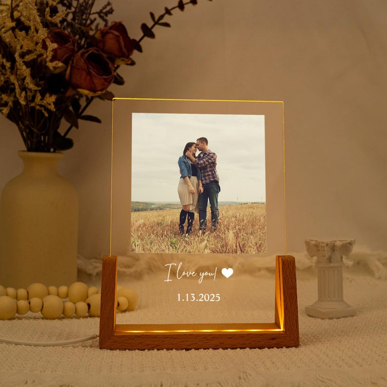 May include: A wooden-based illuminated photo frame. The frame shows a couple in a field, with the text "I love you!" and a heart symbol, plus the date 1.13.2025. The frame emits a warm light.