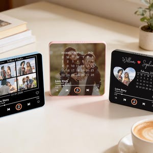 May include: Three square digital photo frames in blue, pink, and black. Each displays a couple's photo, a calendar, and music controls. The frames have the text "Love Story" and names. A cup of coffee sits in the foreground.