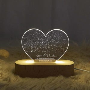 May include: A heart-shaped night light with a constellation design. The light is engraved with the names "Lewis & Caitlin", the date "29.11.2024", and the location "York". The light sits on a wooden base.