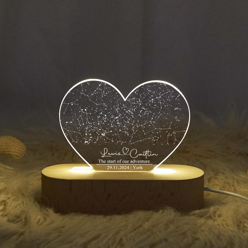 May include: A heart-shaped night light with a constellation design. The light is engraved with the names "Lewis & Caitlin", the date "29.11.2024", and the location "York". The light sits on a wooden base.