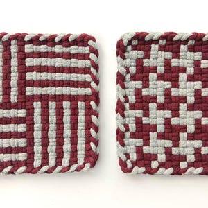May include: Two square potholders made of woven fabric in a maroon and light gray color scheme. The left potholder features a block-striped pattern, while the right potholder displays a repeating houndstooth-style pattern. Both have a braided edge.