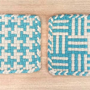 以下が含まれることがあります： 2つの正方形の織り鍋つかみ。各鍋つかみは、青と白の幾何学模様です。端は編み込みの白いトリムで仕上げられています。各鍋つかみの角には小さなループが付いています。