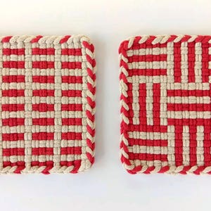 May include: Two square potholders made of woven cotton cord in red and cream. The left potholder features a grid pattern, while the right one displays a geometric block design. Both items have a braided border and a small corner loop.