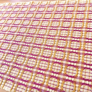 May include: Close-up of a woven textile with a repeating pattern of circles and lines. The design features a combination of magenta, gold, and cream colors. The fabric has a textured appearance, suggesting a handmade or artisanal quality.
