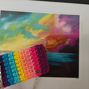 May include: A rectangular crochet handbag featuring horizontal stripes in purple, teal, yellow, peach, and pink. The bag is constructed from thick fabric yarn and includes a silver-toned metal chain strap attached to the sides with metal rings.