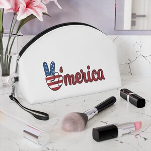 May include: White makeup bag with a black zipper and a black strap. The bag features a peace sign in the colours of the American flag and the word "'merica" in red cursive.