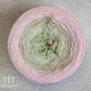 May include: A circular ball of yarn in a gradient of colours, fading from pink to green and cream. A small pink flower is pinned in the centre. The yarn has a subtle shimmer with gold flecks. The logo "Blooming Yarns" is in the bottom left corner.