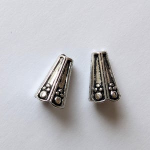 Puede incluir: Dos tapas de cuentas de metal plateado en forma de cono con detalles decorativos. Las tapas tienen un acabado liso y brillante y un diseño ligeramente texturizado.