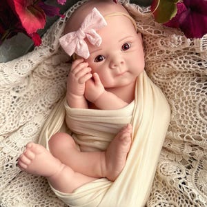 May include: A lifelike baby doll swaddled in a cream-colored blanket, nestled in a cream lace-trimmed basket. The doll wears a pink bow headband. Decorative blue feathers and burgundy flowers surround the baby.