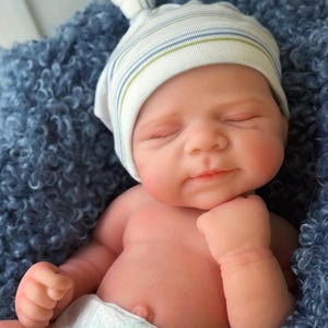 May include: A realistic newborn doll with light skin, closed eyes, and a subtle smile. The doll wears a white knit cap with thin blue and green stripes and a knotted top, along with a white disposable diaper. It rests against a textured blue fabric.