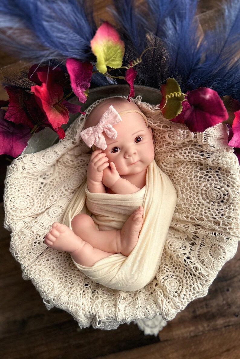 May include: A lifelike baby doll swaddled in a cream-colored blanket, nestled in a cream lace-trimmed basket. The doll wears a pink bow headband. Decorative blue feathers and burgundy flowers surround the baby.