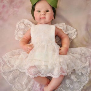 May include: A baby doll dressed in a white lace dress and a green bow headband. The doll has a realistic appearance with rosy cheeks and green eyes. The dress features detailed lace.