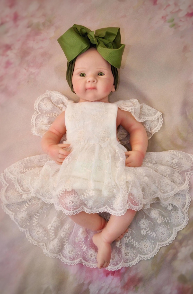 May include: A baby doll dressed in a white lace dress and a green bow headband. The doll has a realistic appearance with rosy cheeks and green eyes. The dress features detailed lace.