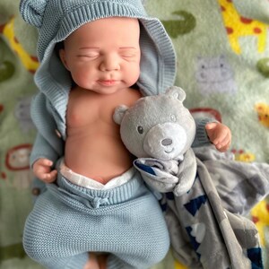 Puede incluir: Una mu&ntilde;eca beb&eacute; vestida con un conjunto de punto azul claro con orejas de oso en la capucha. La mu&ntilde;eca sostiene un peluche de oso gris y est&aacute; acostada sobre una manta verde con estampados de animales.