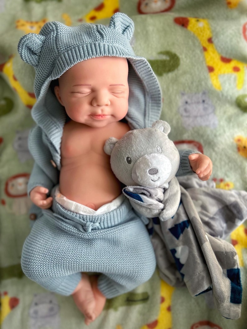 Puede incluir: Una mu&ntilde;eca beb&eacute; vestida con un conjunto de punto azul claro con orejas de oso en la capucha. La mu&ntilde;eca sostiene un peluche de oso gris y est&aacute; acostada sobre una manta verde con estampados de animales.