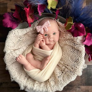 May include: A newborn baby doll wrapped in a cream-coloured swaddle, lying on a cream lace blanket. The doll wears a pink bow headband and is surrounded by purple, red, and green floral accents. The background is a wooden surface.
