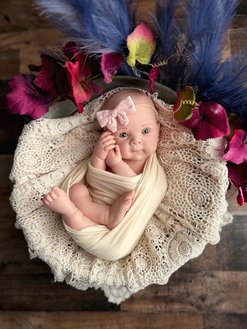 May include: A newborn baby doll wrapped in a cream-coloured swaddle, lying on a cream lace blanket. The doll wears a pink bow headband and is surrounded by purple, red, and green floral accents. The background is a wooden surface.