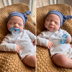 Puede incluir: Una mu&ntilde;eca beb&eacute; realista con gorro azul y body blanco. La mu&ntilde;eca tiene un chupete azul en la boca y est&aacute; acostada sobre una manta de punto marr&oacute;n. Los ojos de la mu&ntilde;eca est&aacute;n cerrados, d&aacute;ndole una apariencia tranquila.
