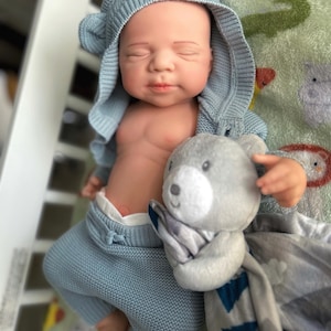 Puede incluir: Una mu&ntilde;eca beb&eacute; realista vestida con un conjunto de punto azul claro, que incluye una chaqueta con capucha con orejas de oso. La mu&ntilde;eca sostiene un osito de peluche gris y una manta estampada gris y azul. El beb&eacute; est&aacute; acostado sobre una manta estampada verde y blanca.