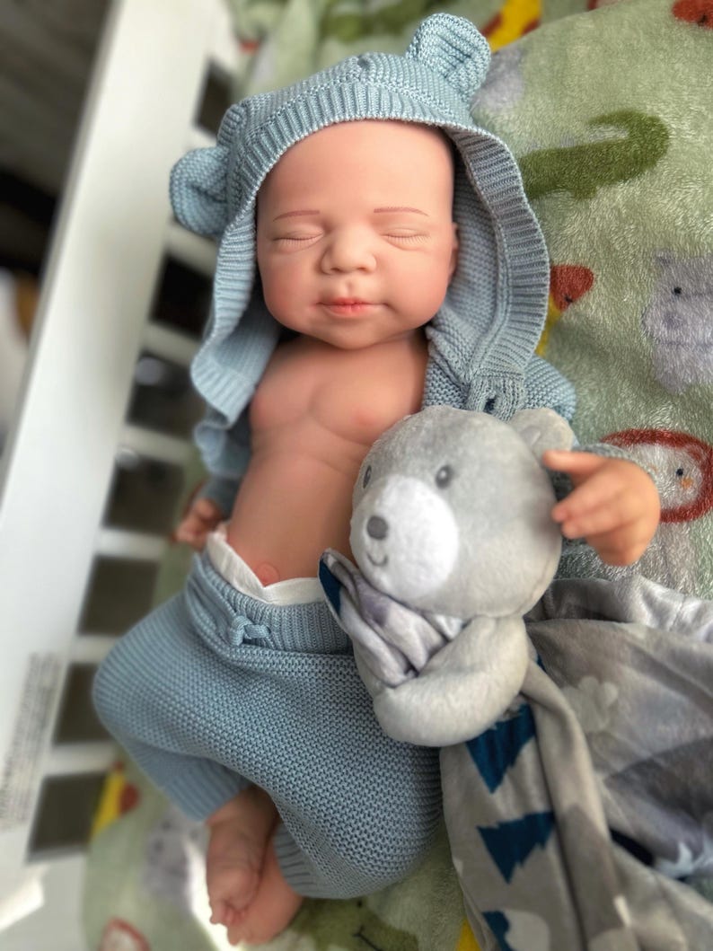 Puede incluir: Una mu&ntilde;eca beb&eacute; realista vestida con un conjunto de punto azul claro, que incluye una chaqueta con capucha con orejas de oso. La mu&ntilde;eca sostiene un osito de peluche gris y una manta estampada gris y azul. El beb&eacute; est&aacute; acostado sobre una manta estampada verde y blanca.