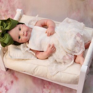 May include: A baby doll wearing a white lace dress and a large green bow, lying on a small white bed with a cream-coloured cushion. The background is a soft pink floral pattern.