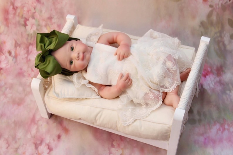 May include: A baby doll wearing a white lace dress and a large green bow, lying on a small white bed with a cream-coloured cushion. The background is a soft pink floral pattern.