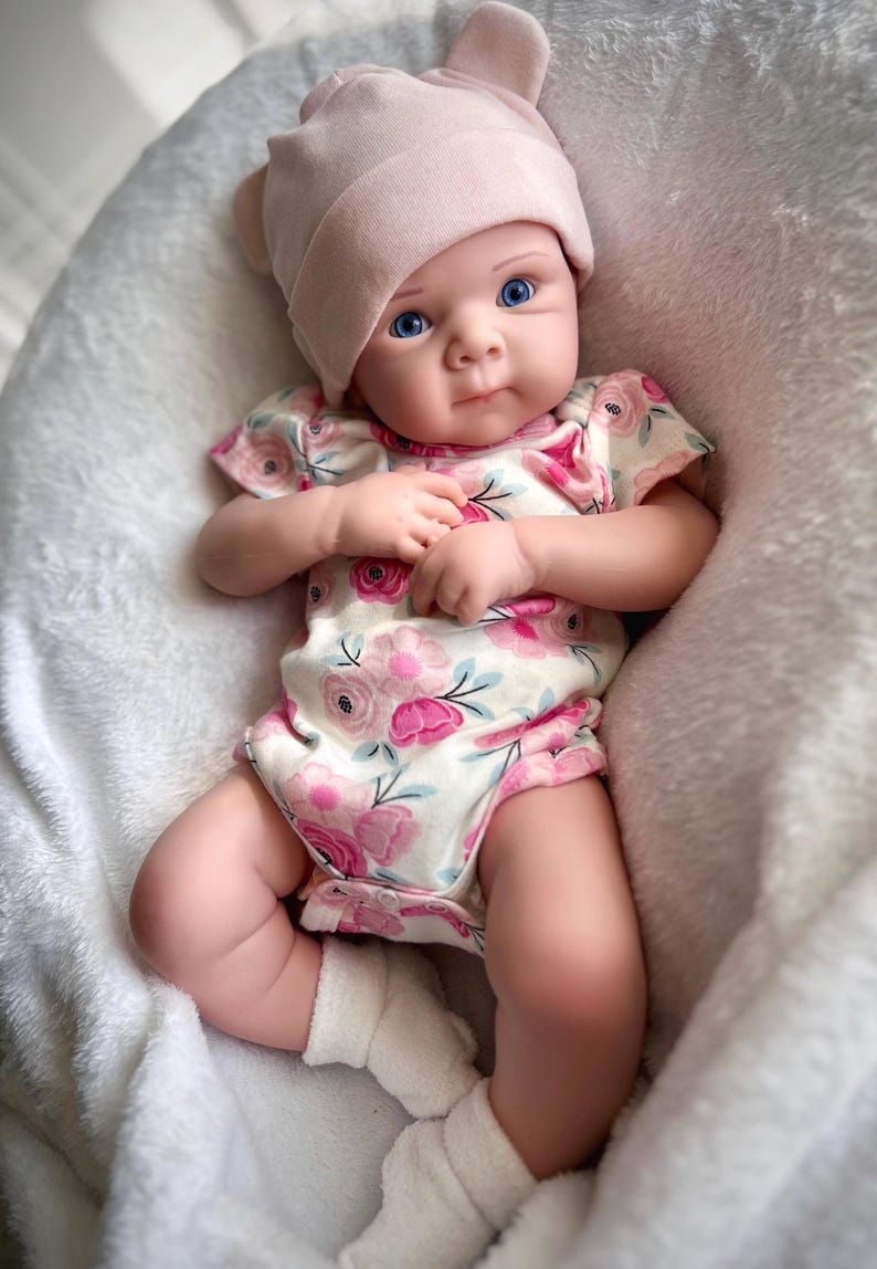 May include: A baby doll with blue eyes, wearing a pink floral print onesie and a matching pink hat with bear ears. The doll is lying on a white, fluffy blanket and wearing white socks.