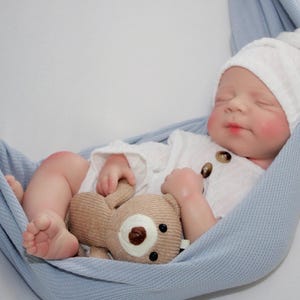 Puede incluir: Una mu&ntilde;eca beb&eacute; realista vestida con un conjunto blanco y un gorro de punto a juego con un pomp&oacute;n, descansando en una hamaca azul claro. La mu&ntilde;eca sostiene un peque&ntilde;o osito de peluche beige. El beb&eacute; tiene mejillas y labios rosados.