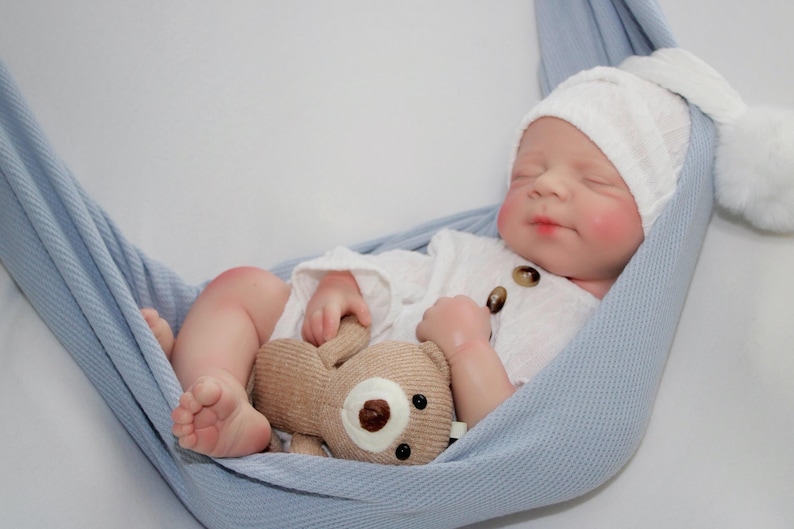 Puede incluir: Una mu&ntilde;eca beb&eacute; realista vestida con un conjunto blanco y un gorro de punto a juego con un pomp&oacute;n, descansando en una hamaca azul claro. La mu&ntilde;eca sostiene un peque&ntilde;o osito de peluche beige. El beb&eacute; tiene mejillas y labios rosados.