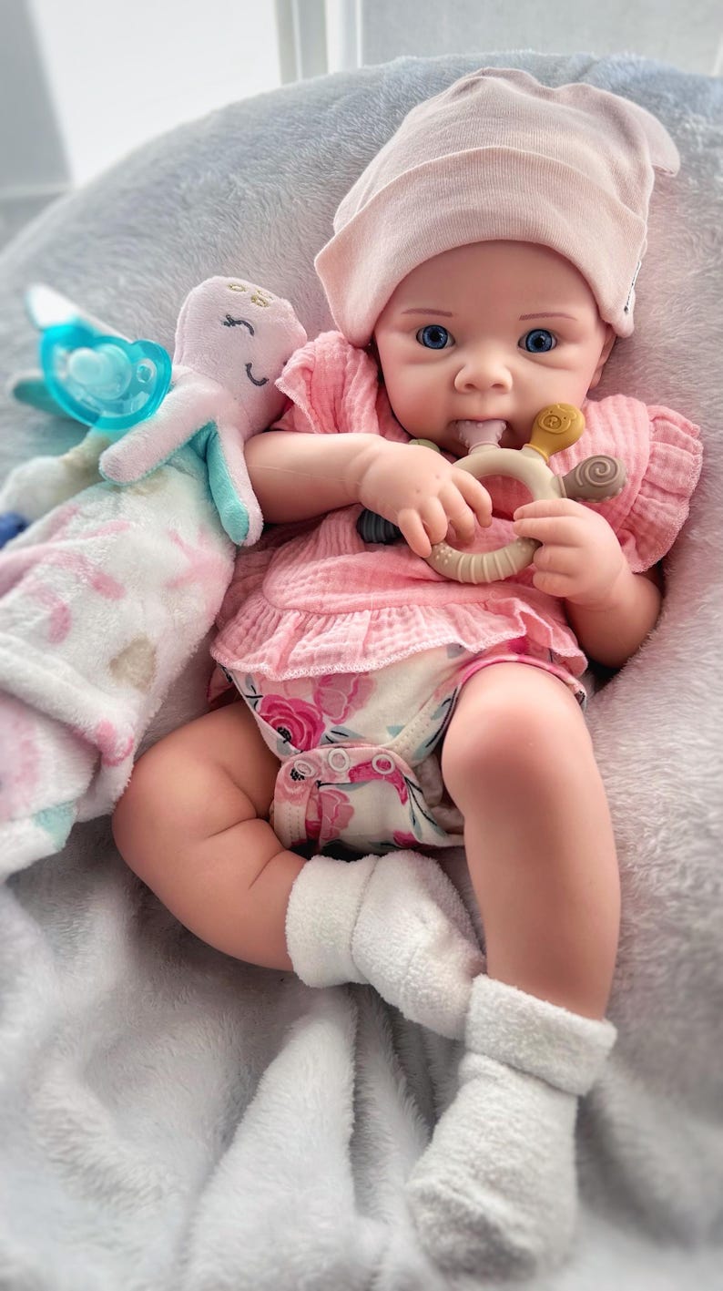 May include: A baby doll wearing a pink floral print onesie, a pink beanie, and white socks. The doll is holding a teething ring in its hand.