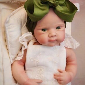 May include: A realistic baby doll with green eyes and a large olive green bow. The doll is dressed in a white dress with lace detailing. The doll is in a white cradle.