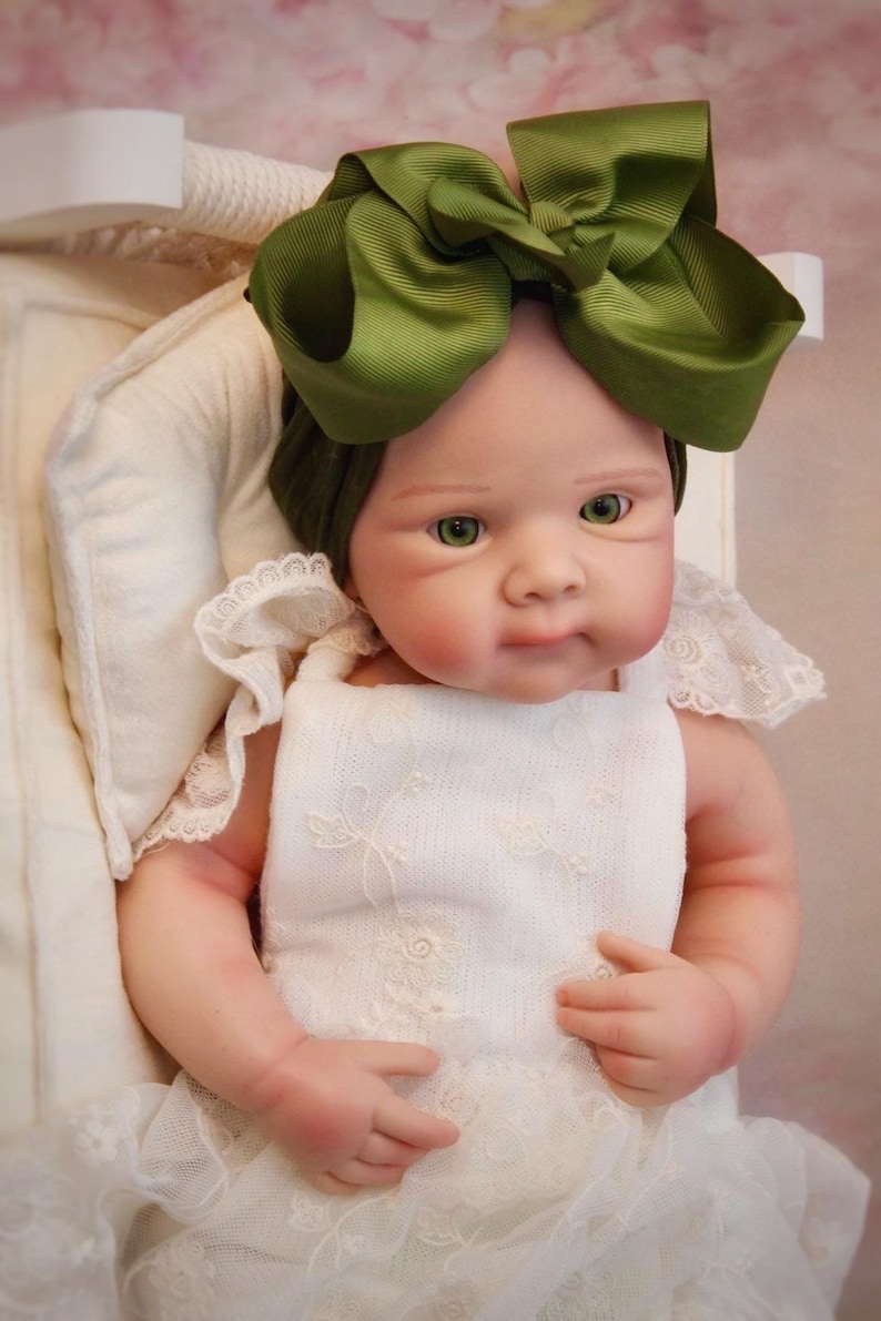 May include: A realistic baby doll with green eyes and a large olive green bow. The doll is dressed in a white dress with lace detailing. The doll is in a white cradle.