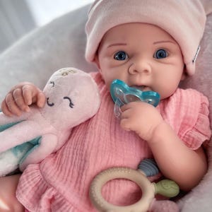 May include: A baby doll with blue eyes, wearing a pink hat and dress, holding a blue pacifier and a pink octopus plush toy. A beige and colorful teether is also visible. The doll is in a soft, light-coloured setting.