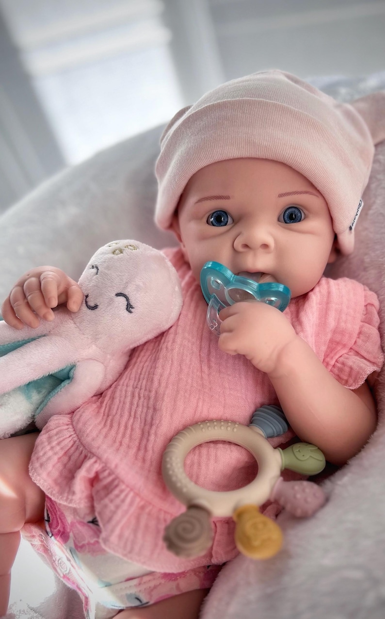 May include: A baby doll with blue eyes, wearing a pink hat and dress, holding a blue pacifier and a pink octopus plush toy. A beige and colorful teether is also visible. The doll is in a soft, light-coloured setting.