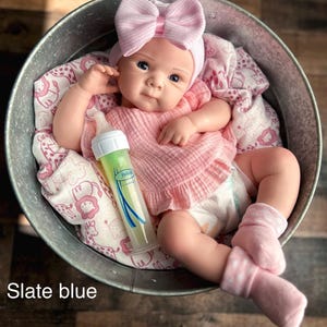 May include: A baby doll in a metal tub, wearing a pink bow headband, pink shirt, and socks. A baby bottle with green liquid is beside the doll. The doll rests on a pink and white patterned blanket. The text "Slate blue" is visible.