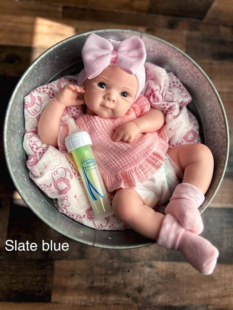 May include: A baby doll in a metal tub, wearing a pink bow headband, pink shirt, and socks. A baby bottle with green liquid is beside the doll. The doll rests on a pink and white patterned blanket. The text "Slate blue" is visible.