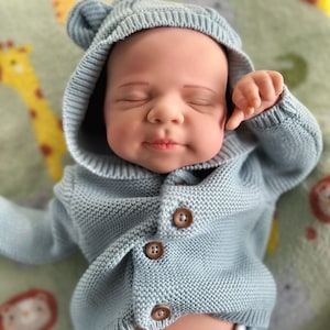 Puede incluir: Una mu&ntilde;eca beb&eacute; con un c&aacute;rdigan de punto azul claro con capucha y orejas de oso. El c&aacute;rdigan tiene tres botones marrones. La mu&ntilde;eca est&aacute; sobre una manta estampada con figuras de animales.