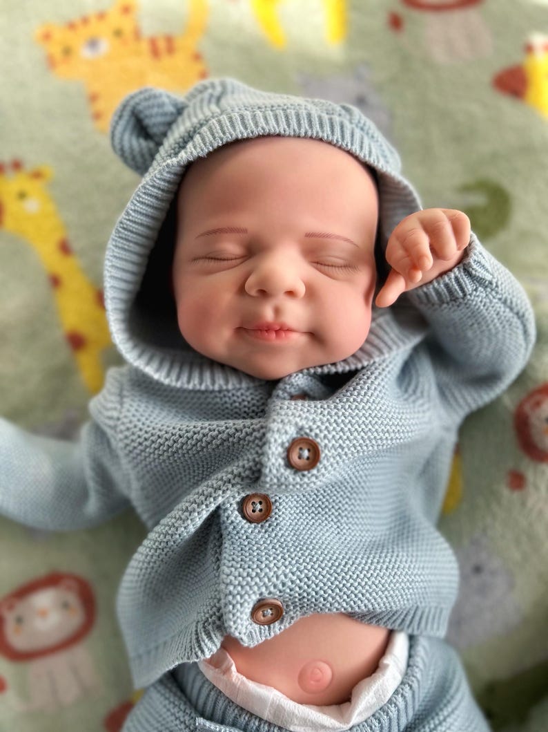 Puede incluir: Una mu&ntilde;eca beb&eacute; con un c&aacute;rdigan de punto azul claro con capucha y orejas de oso. El c&aacute;rdigan tiene tres botones marrones. La mu&ntilde;eca est&aacute; sobre una manta estampada con figuras de animales.