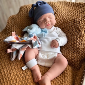Puede incluir: Una mu&ntilde;eca beb&eacute; con gorro de punto azul marino y camiseta blanca de manga larga, tumbada sobre una manta marr&oacute;n. La mu&ntilde;eca sostiene un peluche de elefante azul claro y una manta con etiquetas de colores. El beb&eacute; lleva un pa&ntilde;al y una banda blanca en la pierna.