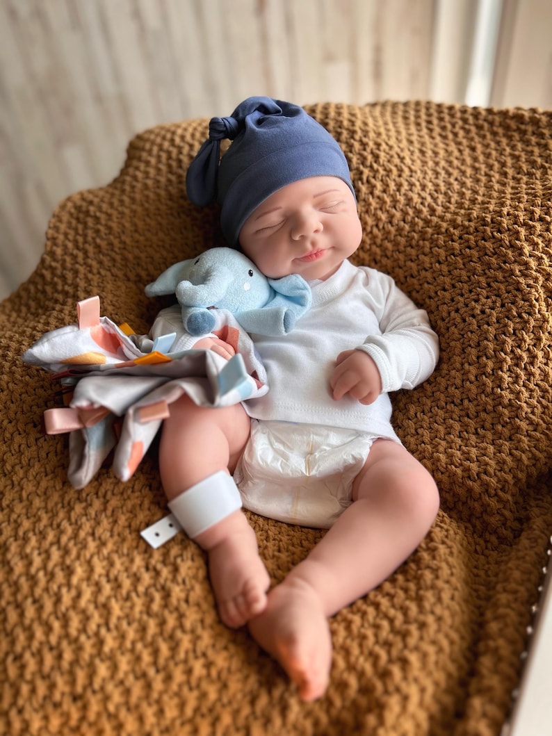 Puede incluir: Una mu&ntilde;eca beb&eacute; con gorro de punto azul marino y camiseta blanca de manga larga, tumbada sobre una manta marr&oacute;n. La mu&ntilde;eca sostiene un peluche de elefante azul claro y una manta con etiquetas de colores. El beb&eacute; lleva un pa&ntilde;al y una banda blanca en la pierna.