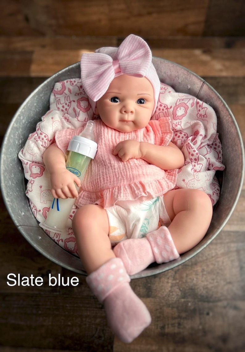 May include: A baby doll in a metal basin, wearing a pink outfit with a bow headband and socks. The doll holds a bottle. The outfit includes a ruffled top. The text "Slate blue" is visible.