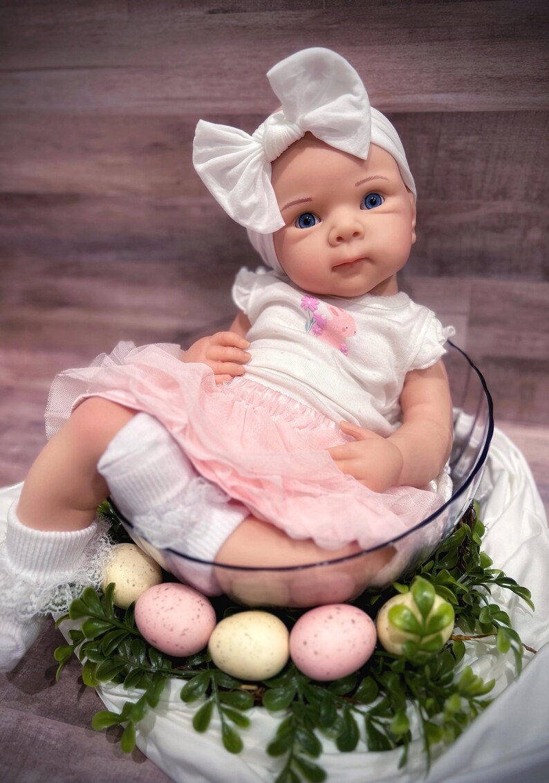May include: A baby doll wearing a white headband with a bow, a white shirt with a pink pig design, a pink tulle skirt, and white socks. The doll is sitting in a clear bowl with green leaves and pink and white speckled eggs around it.