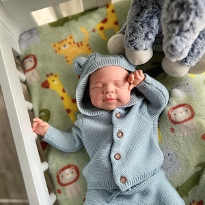Puede incluir: Un beb&eacute; con un conjunto de punto azul claro, que incluye una chaqueta con capucha y pantalones a juego, con botones marrones. El beb&eacute; est&aacute; acostado sobre una manta verde con estampados de animales. Un peluche es visible en el fondo.