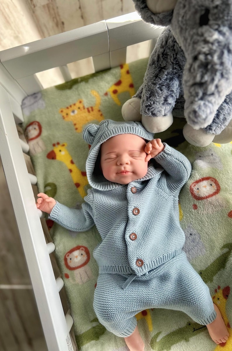 Puede incluir: Un beb&eacute; con un conjunto de punto azul claro, que incluye una chaqueta con capucha y pantalones a juego, con botones marrones. El beb&eacute; est&aacute; acostado sobre una manta verde con estampados de animales. Un peluche es visible en el fondo.