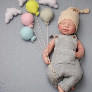 Puede incluir: Una mu&ntilde;eca beb&eacute; reci&eacute;n nacida con un pelele de punto gris y un gorro de punto beige, sosteniendo un manojo de globos de fieltro de colores. Nubes de fieltro blanco est&aacute;n dispersas alrededor de los globos. El beb&eacute; est&aacute; acostado sobre una superficie azul claro.