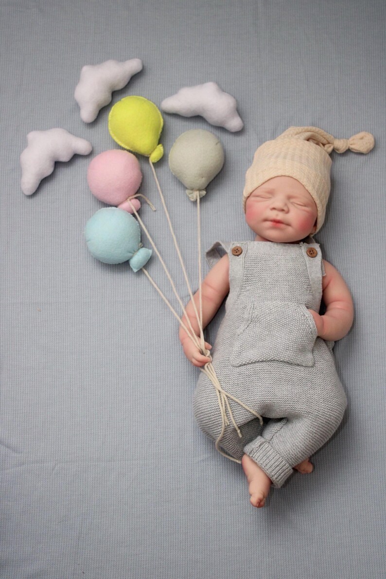 Puede incluir: Una mu&ntilde;eca beb&eacute; reci&eacute;n nacida con un pelele de punto gris y un gorro de punto beige, sosteniendo un manojo de globos de fieltro de colores. Nubes de fieltro blanco est&aacute;n dispersas alrededor de los globos. El beb&eacute; est&aacute; acostado sobre una superficie azul claro.